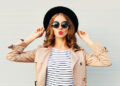 Fashion portrait pretty sweet young woman blowing red lips wearing a black hat sunglasses coat over grey background