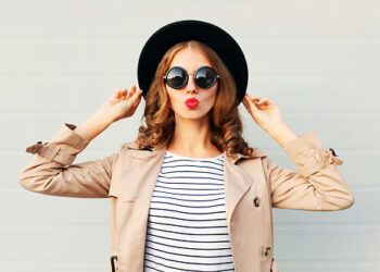 Fashion portrait pretty sweet young woman blowing red lips wearing a black hat sunglasses coat over grey background