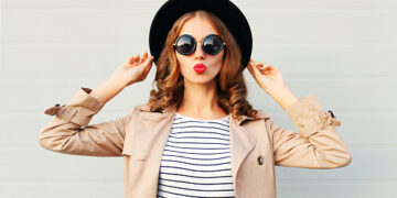 Fashion portrait pretty sweet young woman blowing red lips wearing a black hat sunglasses coat over grey background
